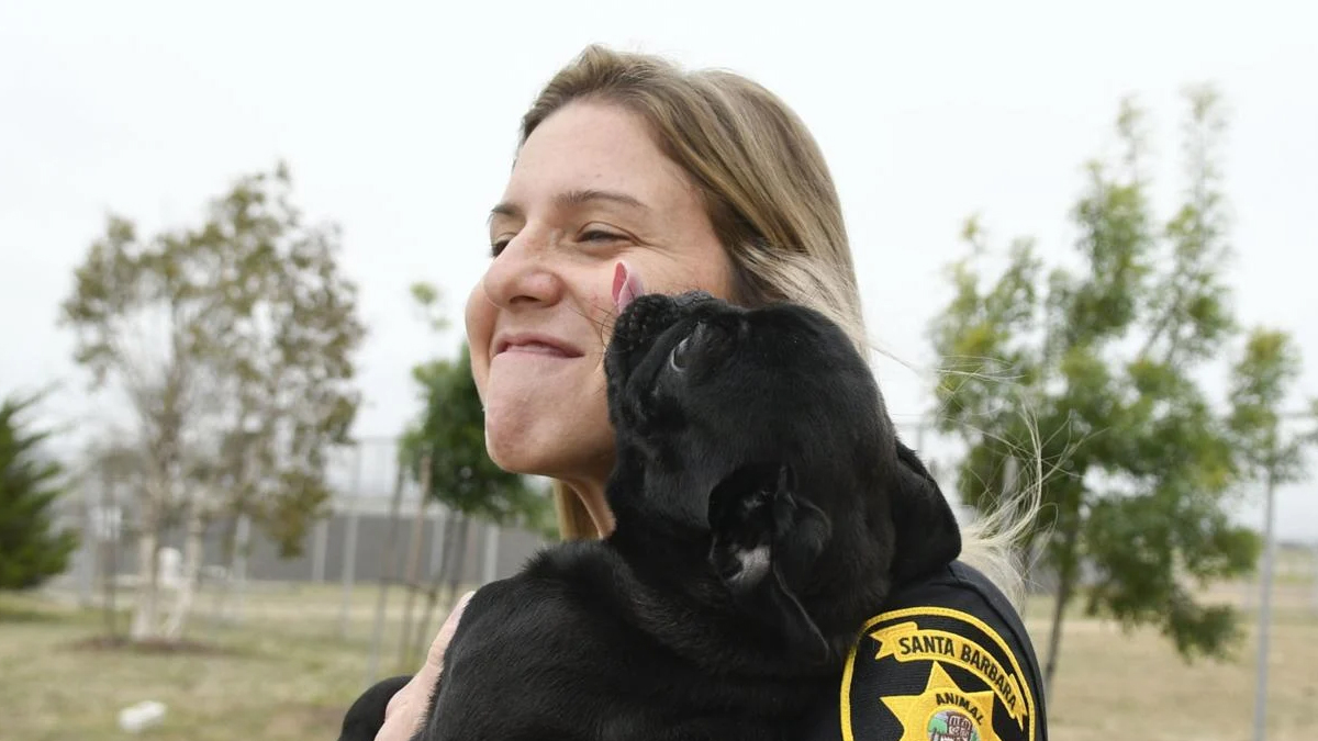 Aubree Gonzales in a park, holding a black dog who is licking her face.