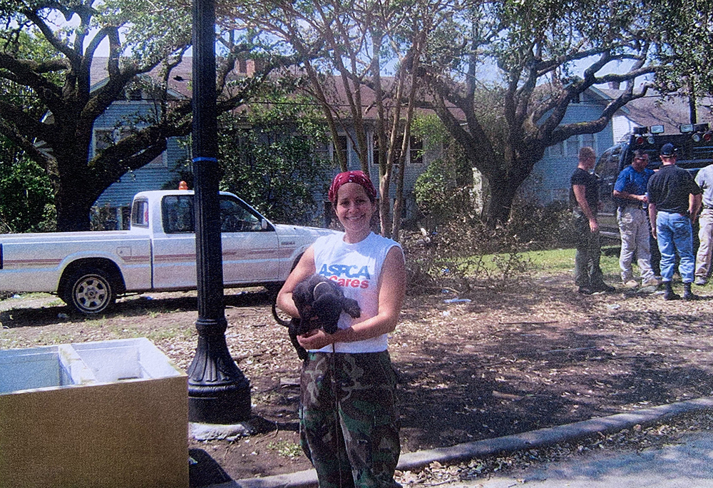 Allison in front of a blue house holding a black dog, wearing an ASPCA tee-shirt and camo pants.