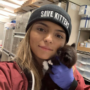 Nadia Oseguera holds a black and white foster kitten close while wearing a hat that says SAVE KITTENS