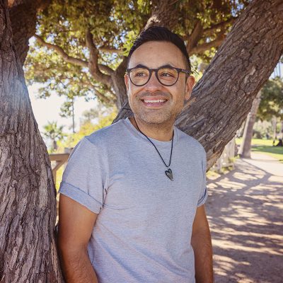 Photo of Jose leaning on a tree in a park, wearing a gray t-shirt and necklace with a heart pendant.